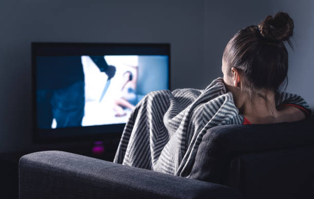 Scary horror movie on tv. Scared woman watching stream service hiding under blanket on couch at night. Sleepless person streaming series or film on television. Alone in dark and afraid of thriller.